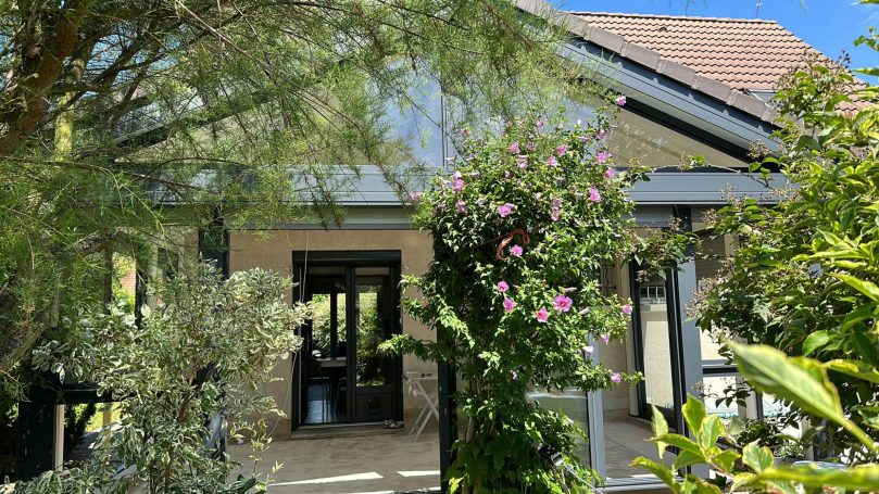 Maison entourée de verdure et de fleurs, avec un toit en pente et de grandes fenêtres.