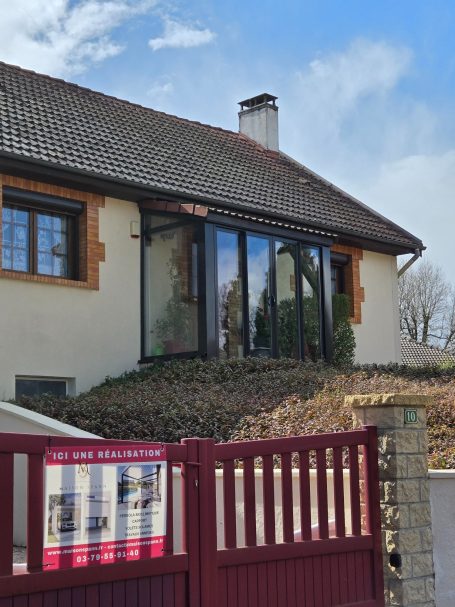 Maison avec véranda, jardin en pente, clôture rouge et ciel bleu.