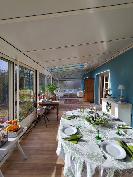 Une véranda lumineuse avec une table dressée et un coin salon en arrière-plan.