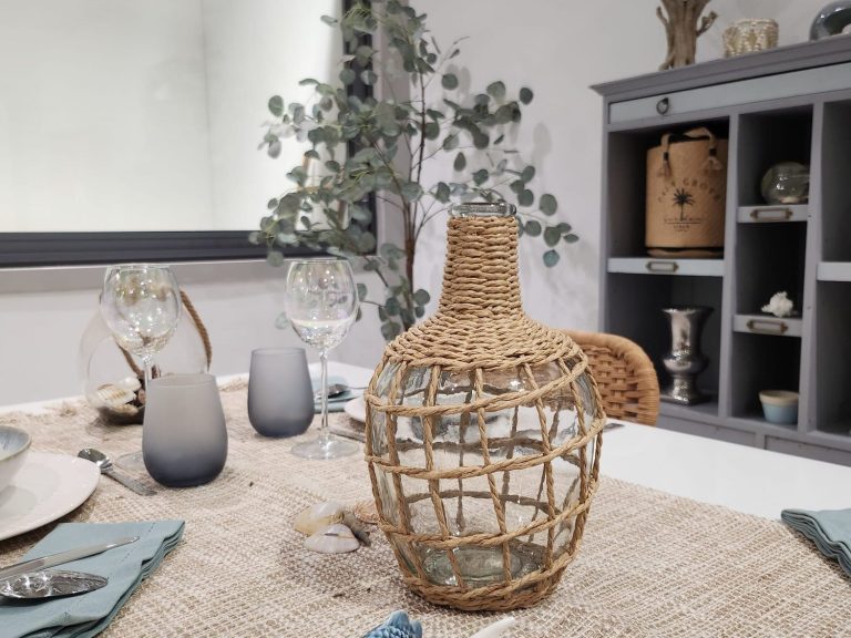 Carafe en osier sur une table décorée, avec des verres et des éléments naturels en arrière-plan.