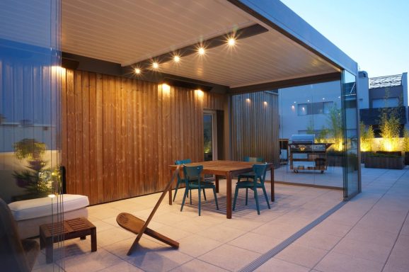 Terrasse moderne avec table, chaises et éclairage, entourée de murs en verre et bois et pergola bioclimatique de la marque Maison Spann