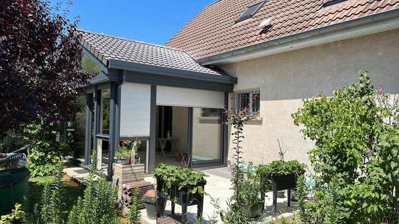 Maison avec une véranda et des plantes en pot, sous un ciel bleu ensoleillé.